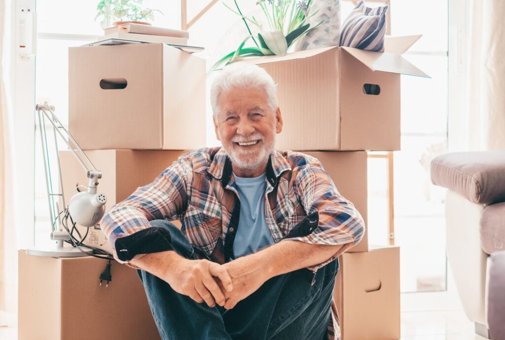 Benefits of moving to senior living early as an older man sits smiling in a bright room surrounded by neatly packed moving boxes, reflecting a confident and well-prepared transition.