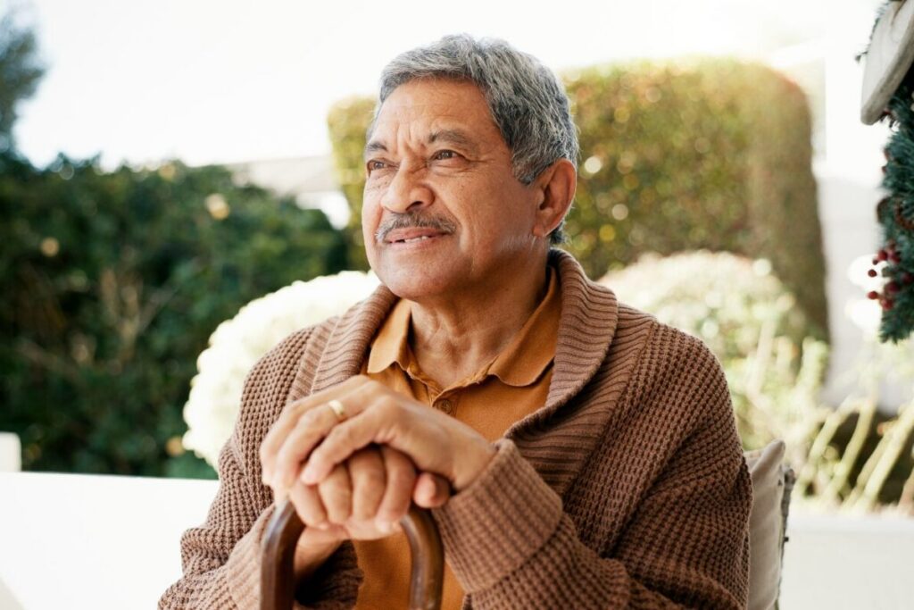 An older man sits peacefully on a sun-filled porch, hands resting on his cane and gaze set on the garden beyond, embodying the quiet confidence and peace of mind that retirement done right can bring.