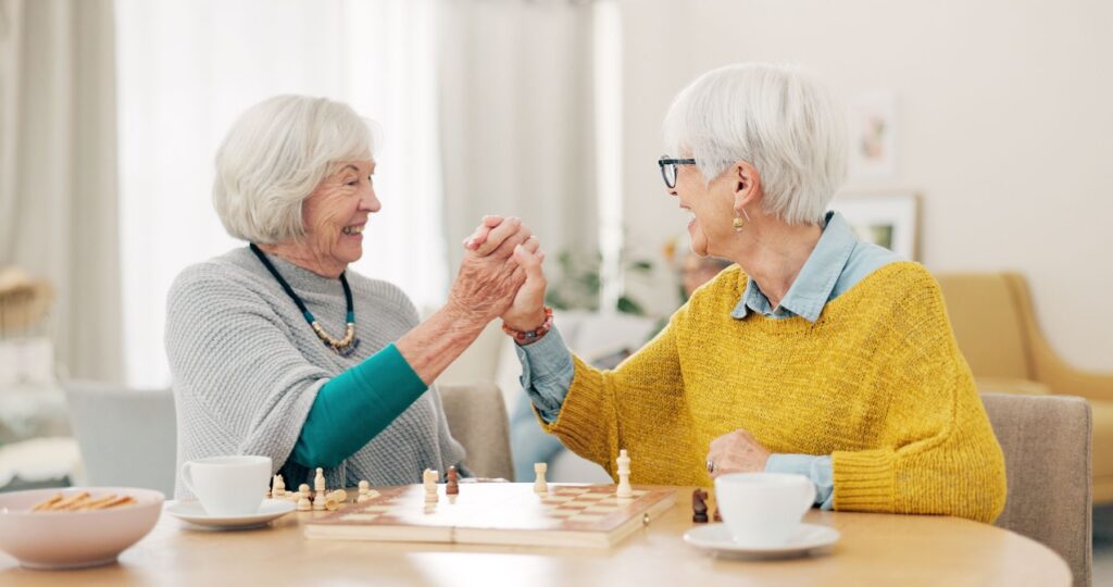 Two older women sit across from each other at a table, laughing and clasping hands mid-game over a chessboard with coffee and snacks nearby — a lighthearted moment that captures the importance of belonging for seniors, built through shared games and genuine friendship.